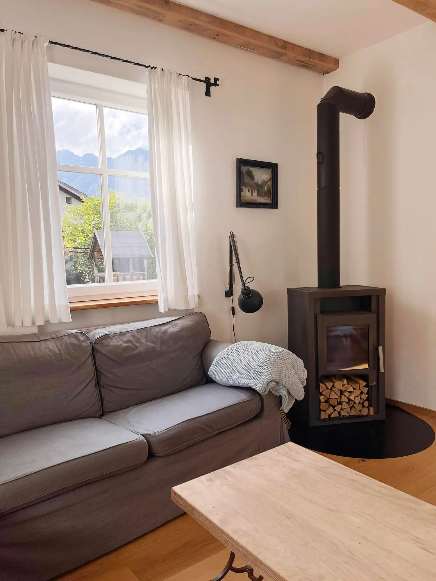 Wohnzimmer mit Sofa und Kaminofen, durchs Fenster die Berge